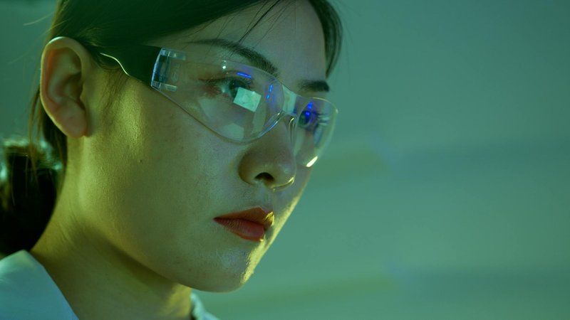 Close-up of a female scientist wearing safety goggles in a lab setting with green lighting.