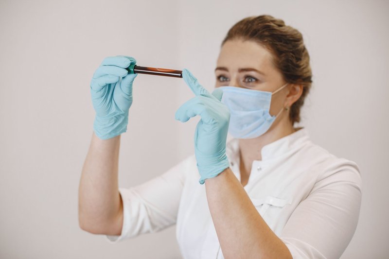 A female healthcare worker wearing gloves and a mask examines a blood sample in a test tube.