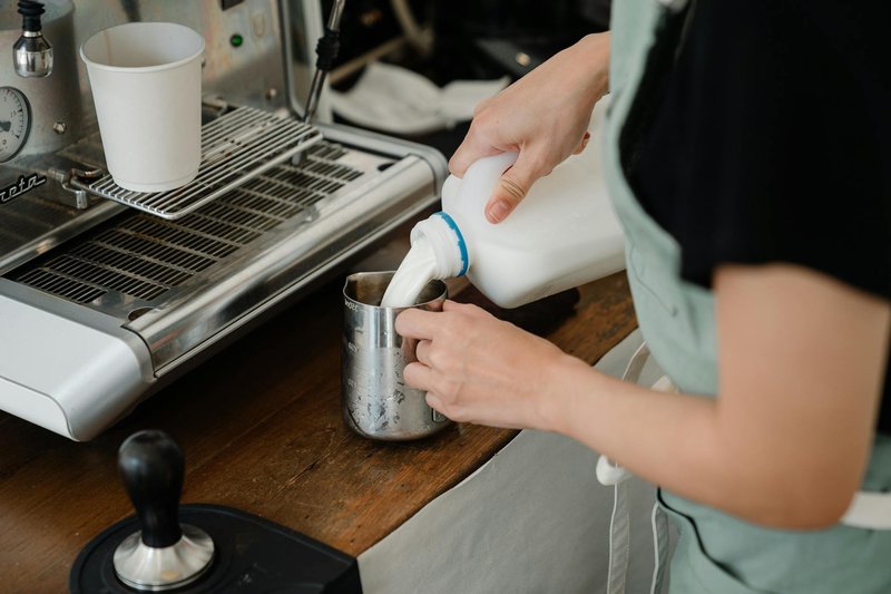 Side view of crop unrecognizable cafe worker pouring fresh milk into big stainless still cup while preparing beverage at counter with coffee machine in modern cafe