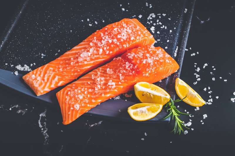 Close-up of raw salmon fillets garnished with salt, lemon wedges, and rosemary on a baking tray.