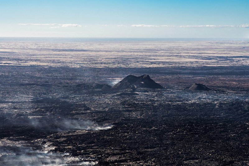 A wide aerial view of a rugged volcanic landscape with visible lava flows and smoke rising from the surface.