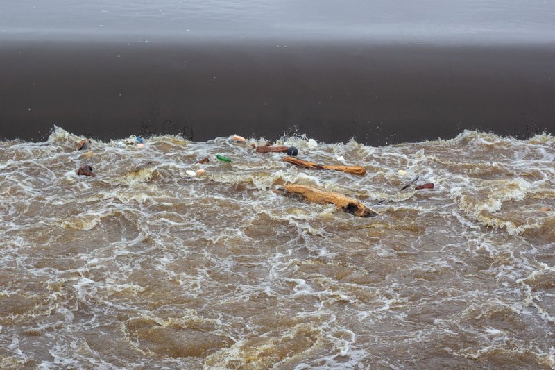 A polluted river scene showing floating trash, wood, and debris in turbulent water.