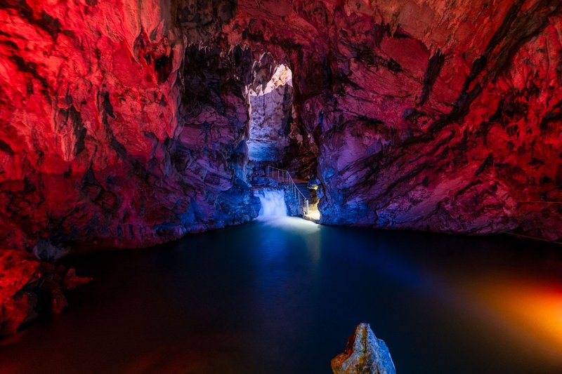 Colorful cave interior with illuminated walls and a waterfall cascading into a serene pool.