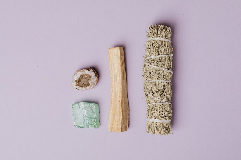 Flat lay of spiritual healing tools including sage, palo santo, quartz geode, and amazonite on a lavender background.