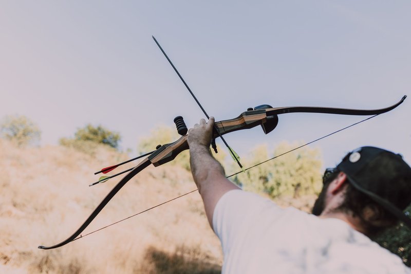 Man aiming a recurve bow outdoors in a forest setting under a blue sky.