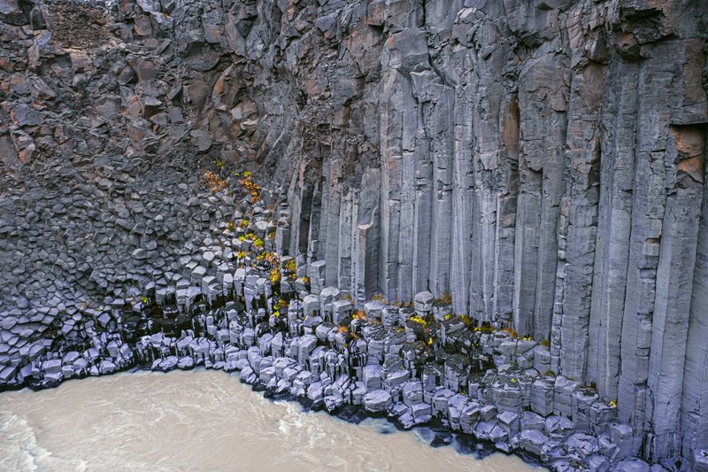 Beautiful eroded basalt columns form a dramatic cliff by a tranquil river with patches of yellow foliage.