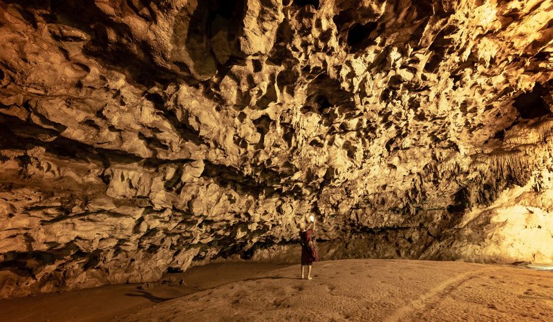 Person exploring large cave with limestone formations, illuminated by warm light.