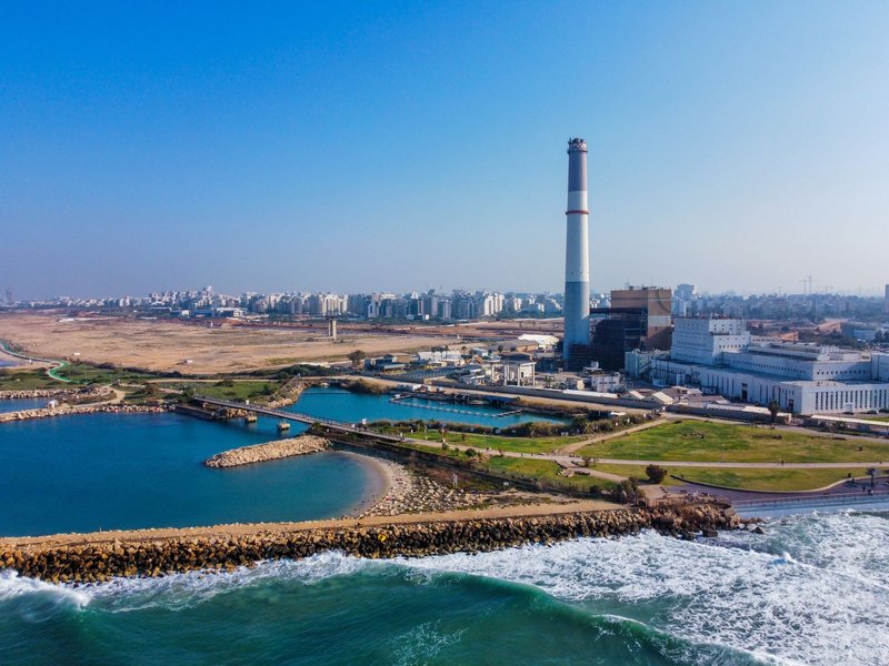 Aerial view of a coastal factory with city skyline, clear sky, and ocean waves.