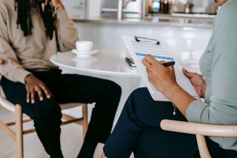 Crop unrecognizable female psychotherapist or interviewer asking questions and taking notes in clipboard during interview with black male