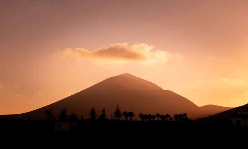 Silhouette of a volcano in Lanzarote at sunset, with a warm orange sky and clouds.