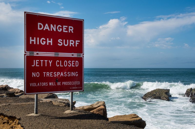 A warning sign for high surf and closed jetty near a rocky seashore with waves crashing.