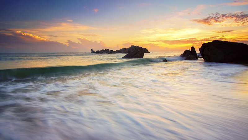 Stunning sunset over a rocky beach with waves and colorful sky, perfect for summer travel inspiration.