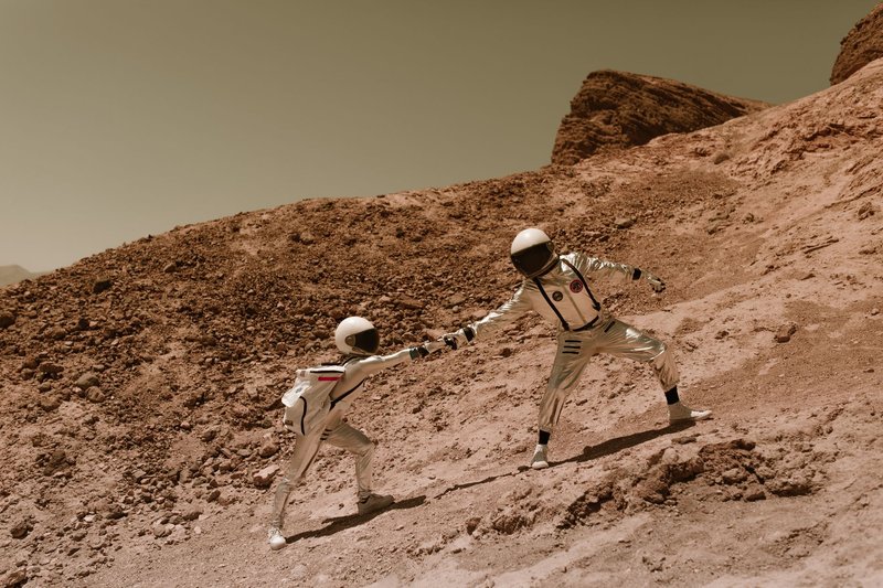 Two astronauts in space suits assisting each other on a rocky terrain resembling Mars.