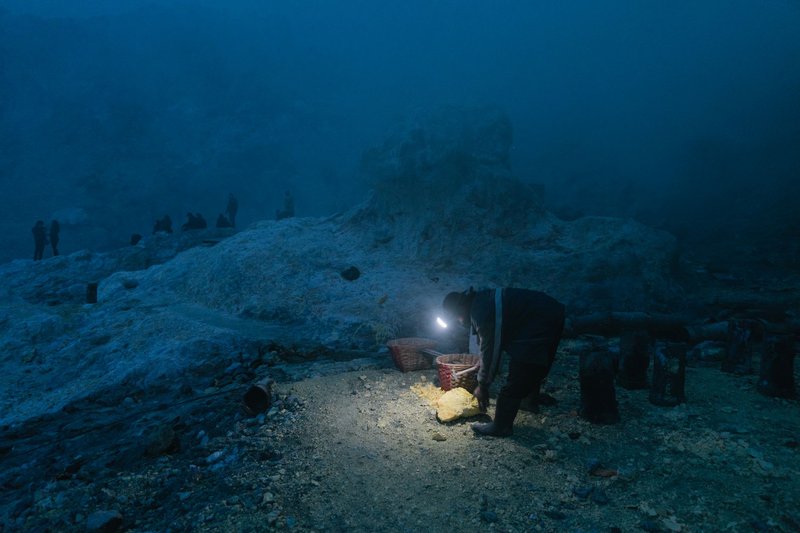 A miner using a flashlight while extracting sulfur in a dark quarry, creating a dramatic nighttime scene.
