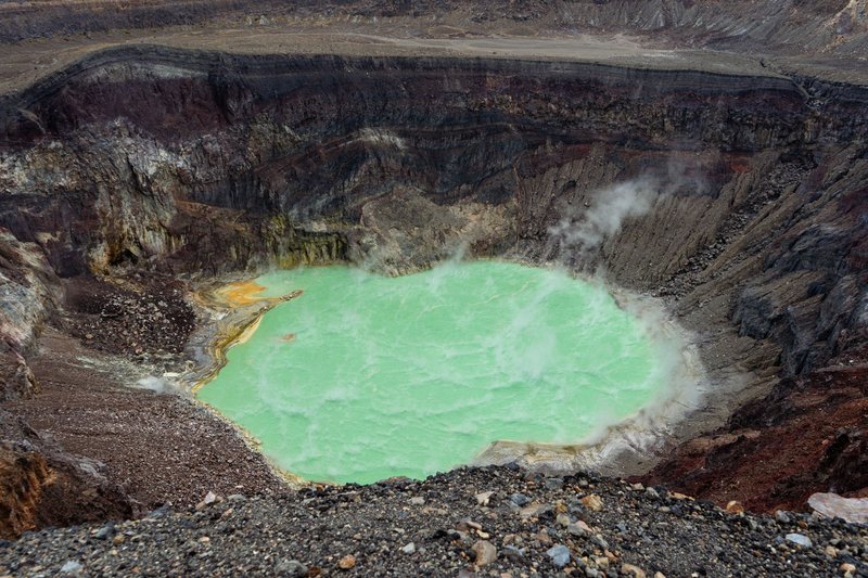 An overhead shot of a steaming volcanic crater with a vibrant green lake, showcasing geothermal activity.