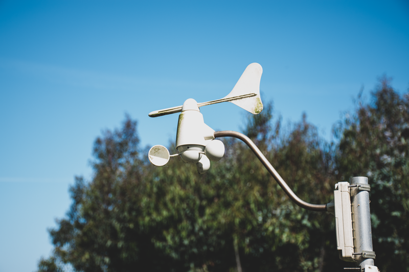 A white anemometer measuring wind speed against a clear blue sky, surrounded by greenery.