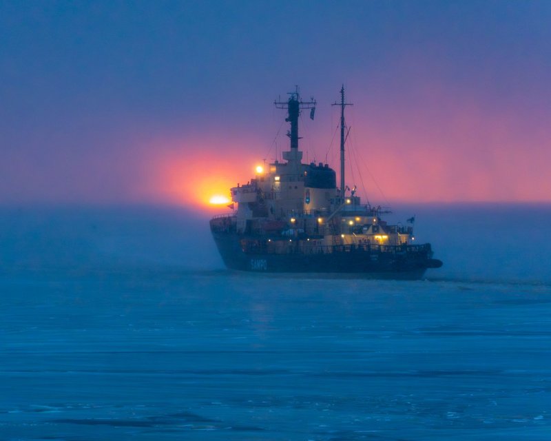 The icebreaker ship Sampo cuts through icy waters during a vibrant sunset, creating a dramatic scene.