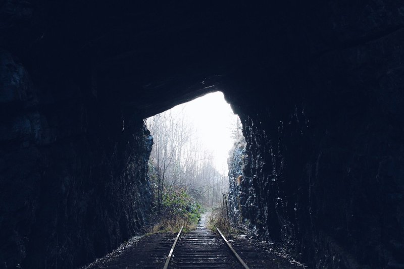 A dark railway tunnel with tracks leading to a bright natural landscape.