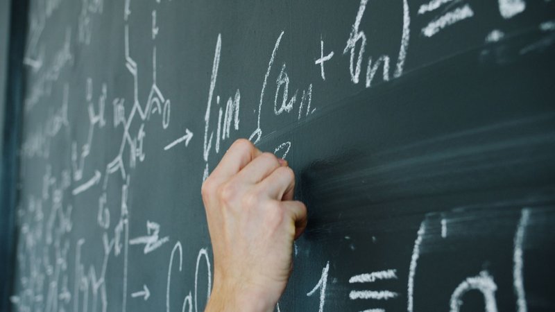 Close-up of a hand writing mathematical equations on a classroom chalkboard.