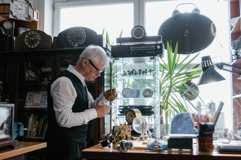 Elderly man repairing clocks in a workshop surrounded by timepieces and tools.