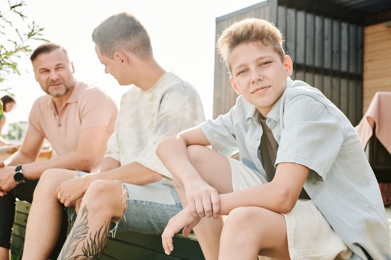 A cheerful boy sits outdoors with family enjoying a sunny day.